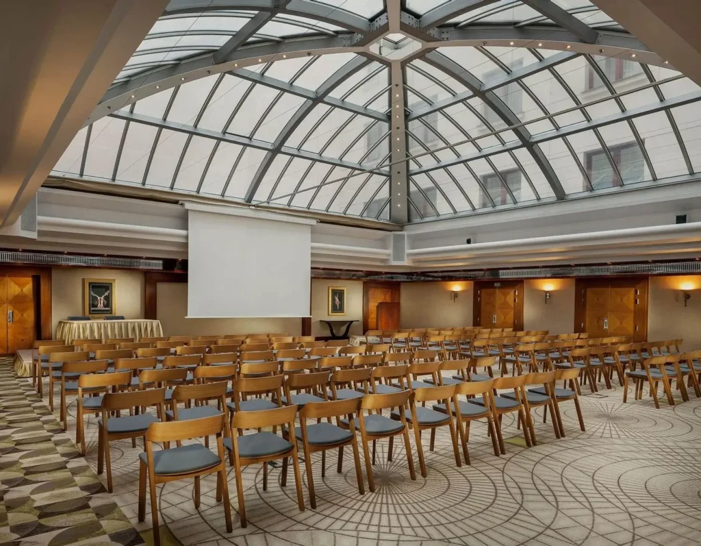 Geräumiger Konferenzraum bei Alcron Prag, mit Reihen von Holzstühlen vor einer Projektionsfläche unter einer großen Glasdecke.